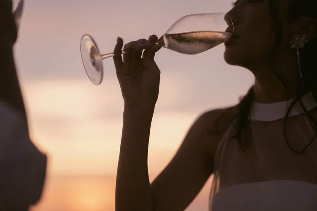 The bride sips champagne in silhouette against a pink and violet sky, a long drop earring at her ear, at sunset on her Miyakojima wedding day.