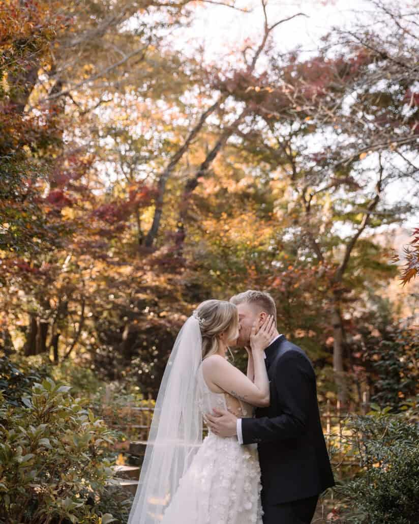 The bride holds the groom's face for a kiss under red and gold maples in a Hadano ryokan garden, an autumn elopement in Japan.