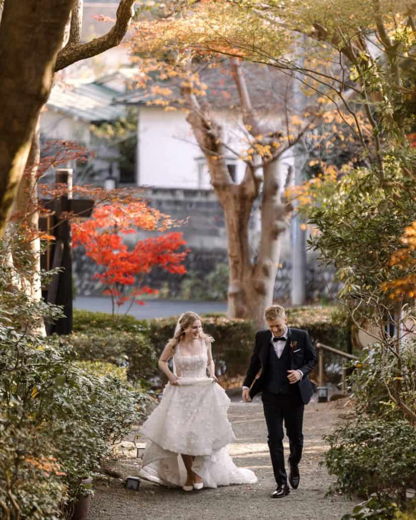 The bride lifts her skirt and walks a gravel garden path with the groom, both laughing, a red maple behind them, at a ryokan in Hadano.