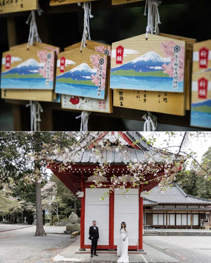 Wooden ema prayer plaques painted with Mt Fuji hang at the shrine, and the couple stand by a red-and-white hall under cherry blossom branches.