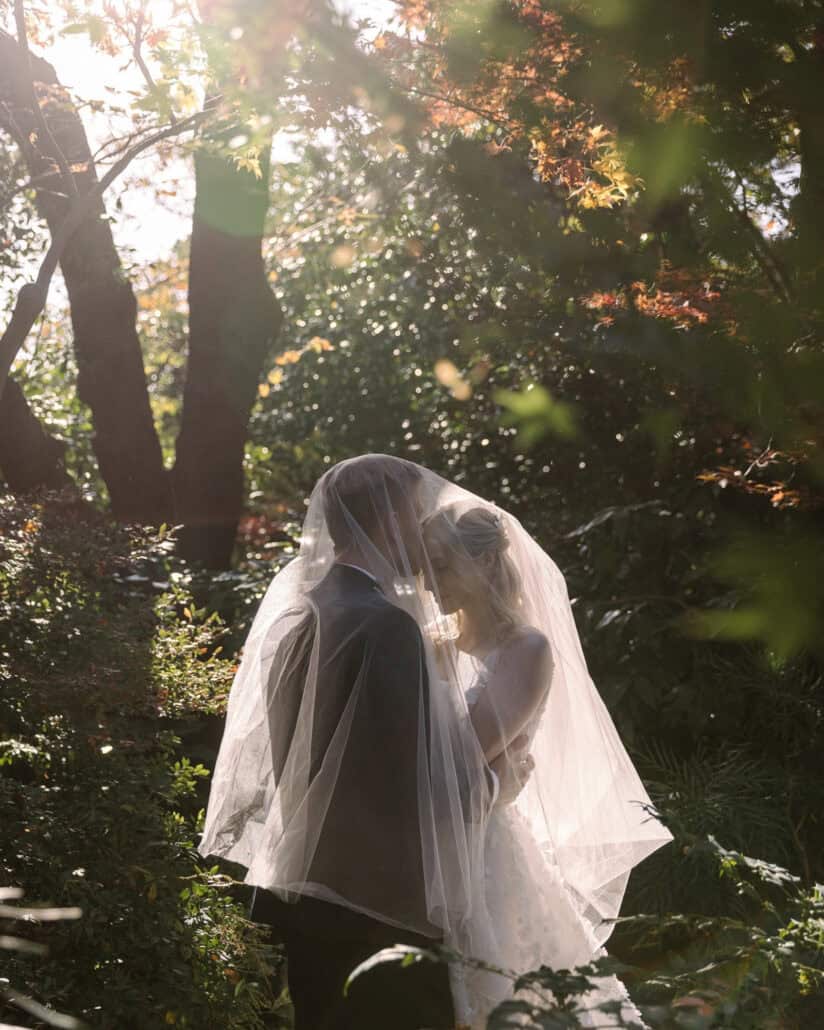 Backlit by autumn sun, the bride and groom stand close under her veil among the maples of a ryokan garden, an autumn elopement in Japan.