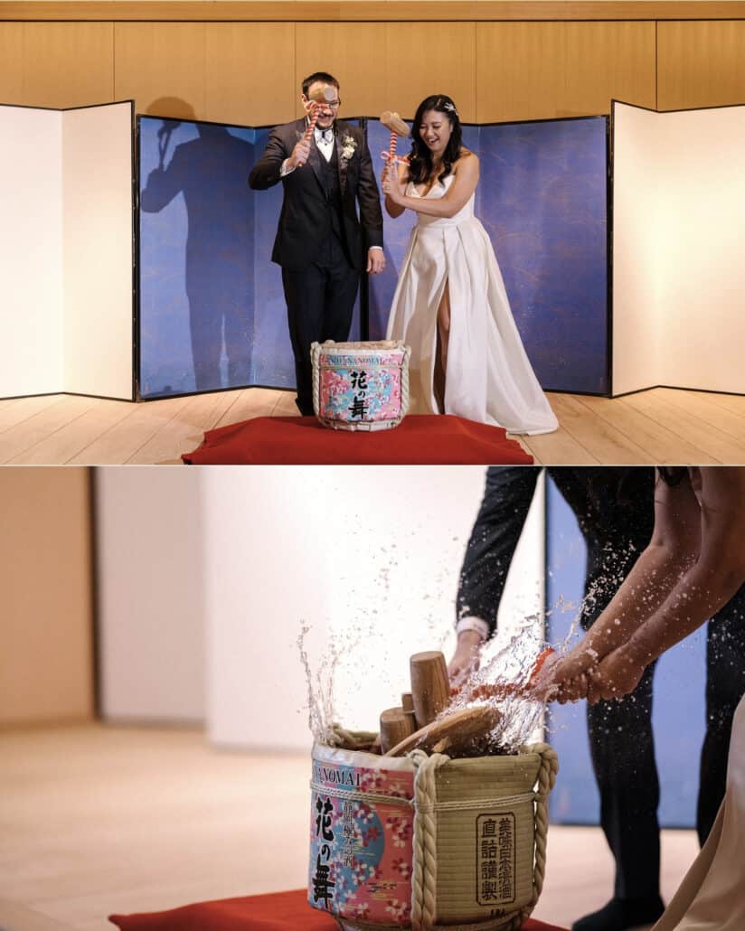 The couple swing wooden mallets at a sake barrel for kagami biraki in front of a blue folding screen, and the sake splashes up from the barrel.