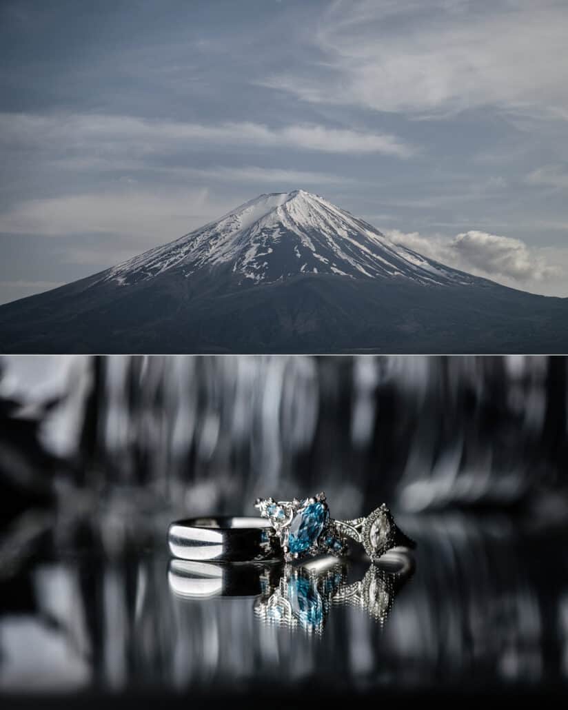Mt Fuji under streaks of cloud, and the wedding rings, a plain band beside rings set with blue stones and a clear stone, on a dark mirrored surface.