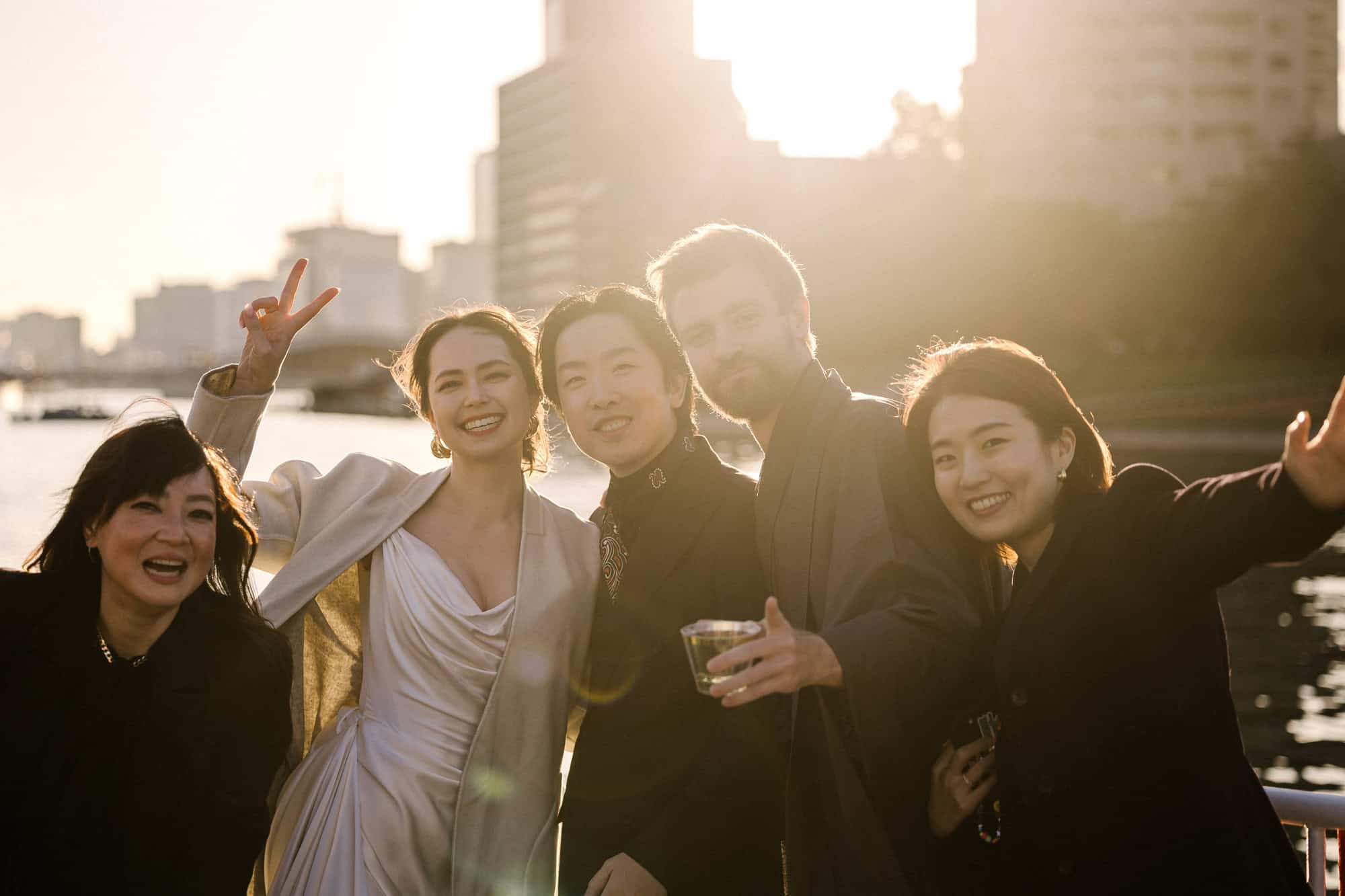 The bride in a cream coat crowds in with laughing friends on a boat deck on Tokyo Bay, low sun behind the towers, on a wedding cocktail cruise.