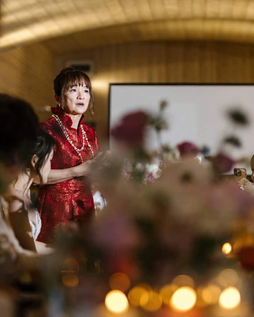 A woman in a red qipao and pearls gives a speech at the wedding dinner at Fujisan Winery, the bride seated beside her and candles glowing in front.