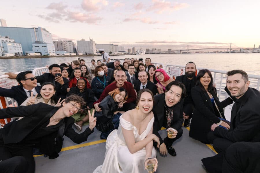 Wedding guests pack the open upper deck around the bride and groom, drinks raised, with Rainbow Bridge and a pink sky over Tokyo Bay behind.