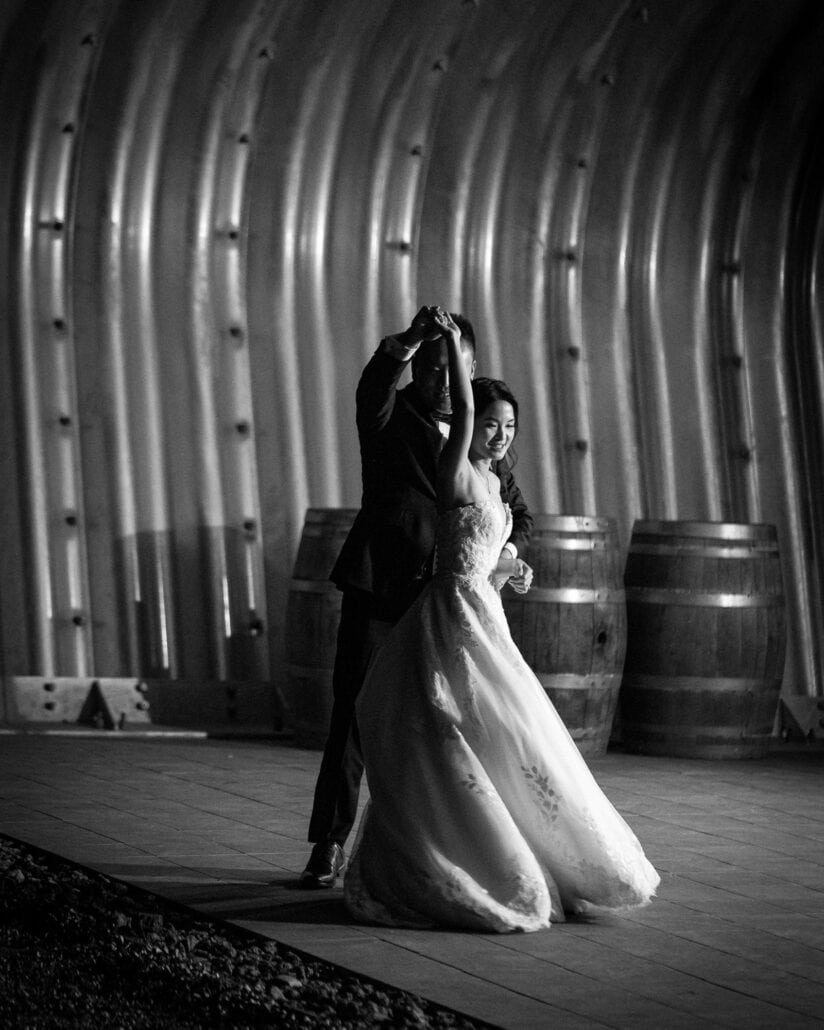 In black and white, the groom twirls the bride in their first dance under a corrugated steel vault lined with wine barrels at Fujisan Winery.