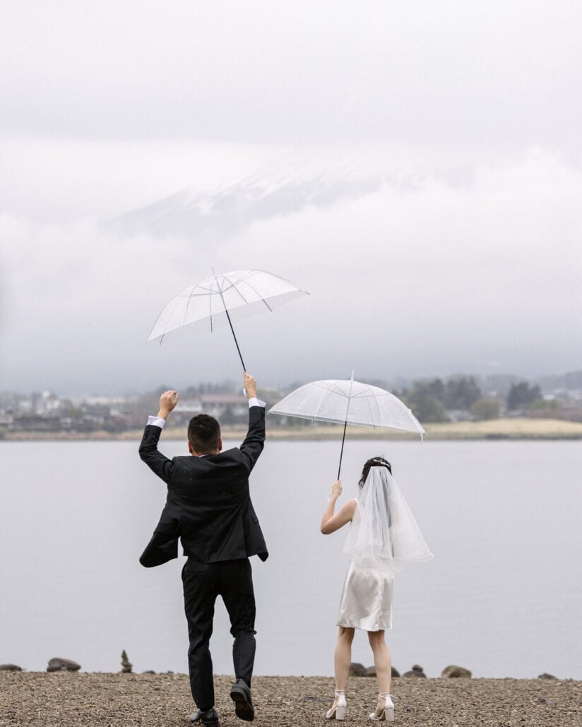 The groom punches the air and the bride lifts a clear umbrella on a pebbled lakeshore the day after the wedding, Mt Fuji's snow faint in the cloud.
