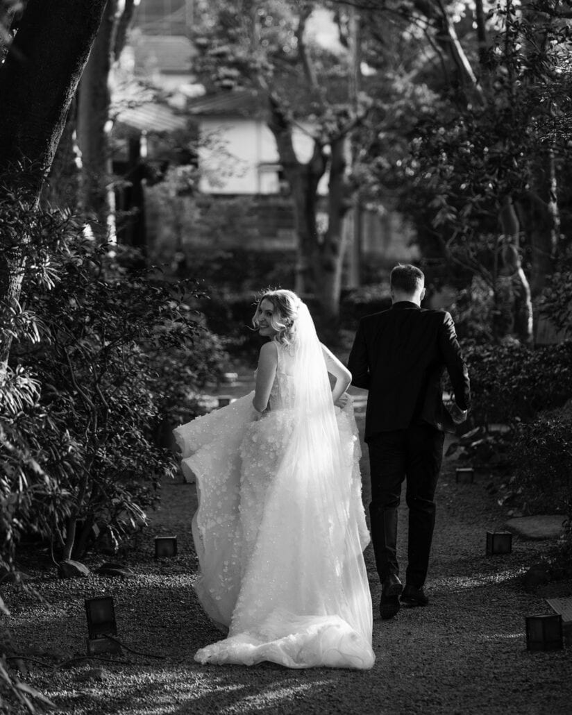 In black and white, the bride looks back, lifting her dress as she follows the groom down a lantern lined path at a ryokan wedding in Japan.