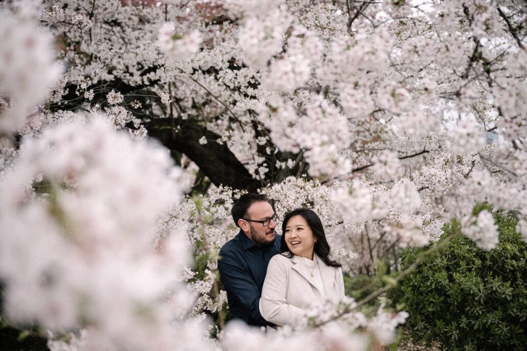 He holds her from behind under a cherry tree in full bloom in a Tokyo park, soft blossoms framing them in the foreground.