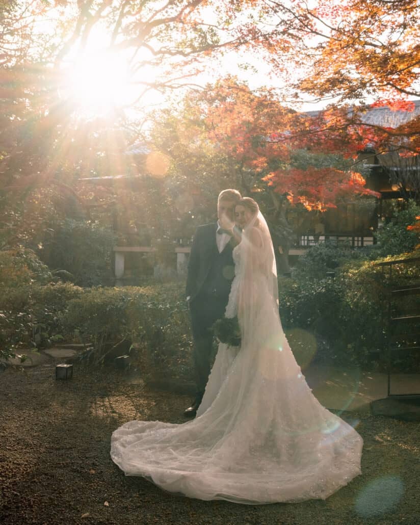 Sunlight flares through red maples as the bride and groom embrace, her long train across the gravel, an autumn elopement in Japan at Jinya.