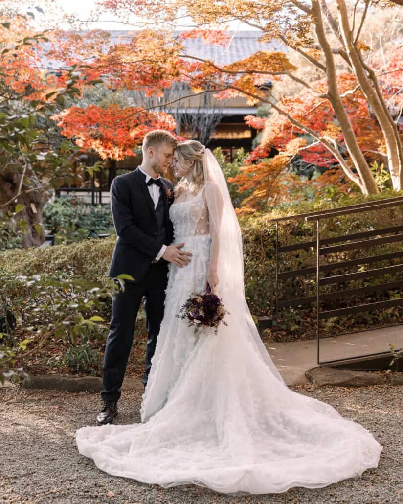 The bride and groom stand forehead to forehead under red maples by a traditional roofline in the garden at Jinya, an autumn elopement in Japan.