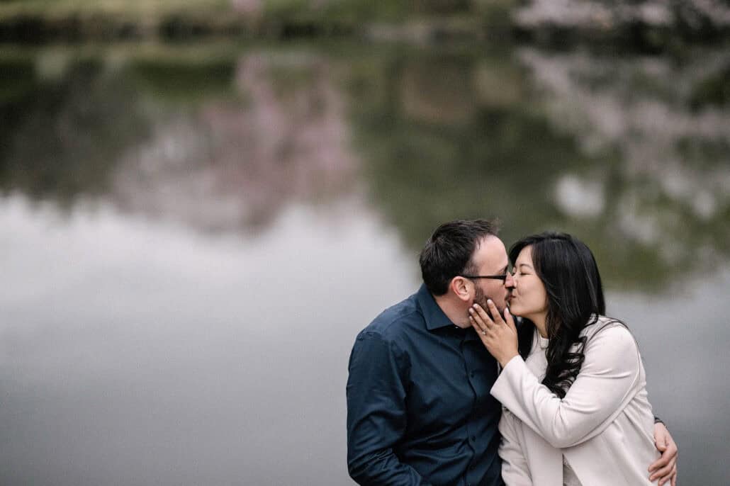 The couple kiss by a pond in a Tokyo park, her hand and engagement ring at his cheek, cherry blossoms reflected in the water behind them.