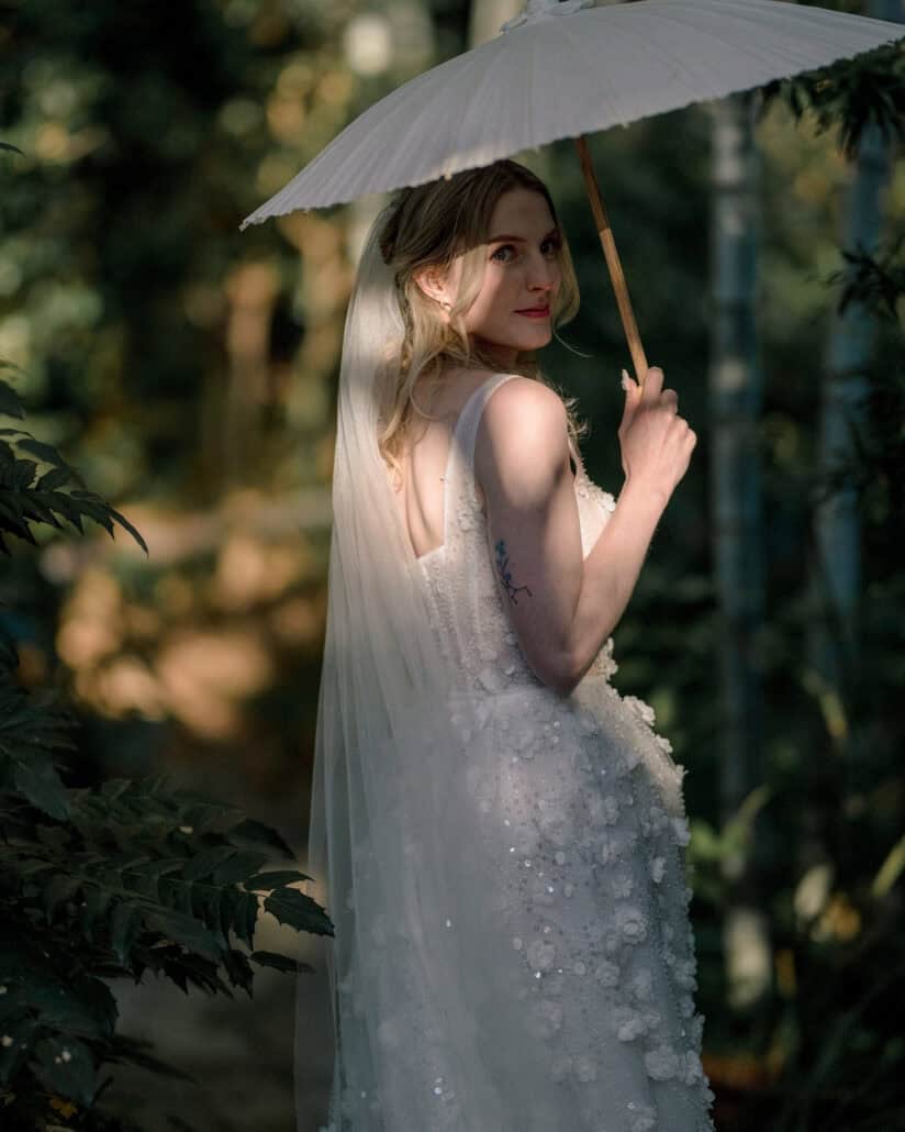 Komorebi, sunlight through the leaves, falls across the bride under a white paper parasol as she looks back over her shoulder on her Japan elopement.