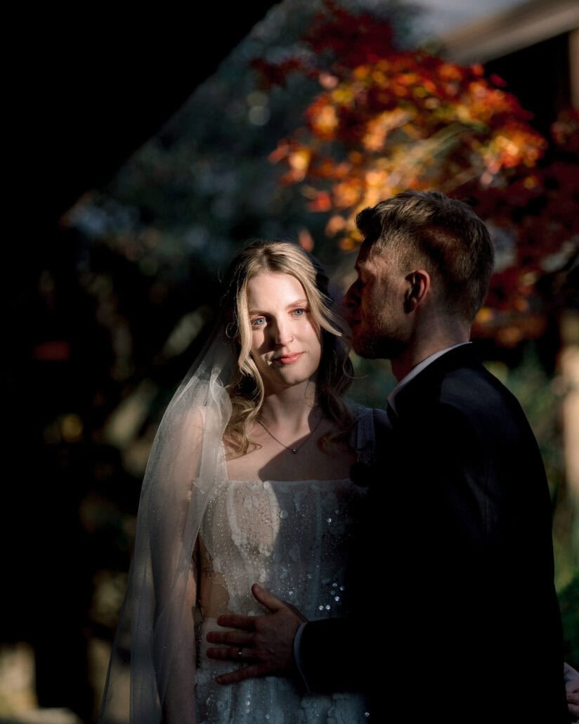 A shaft of light falls on the bride's face as the groom leans into her hair, red maple leaves glowing behind them under the ryokan eaves.