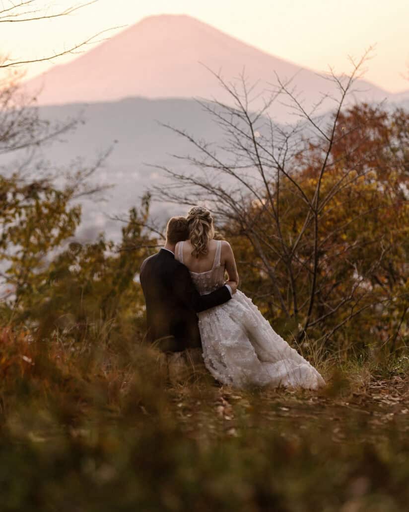 Backs to the camera, the bride and groom sit close on an autumn hillside as Mt Fuji turns pink at sunset beyond the hazy valley.