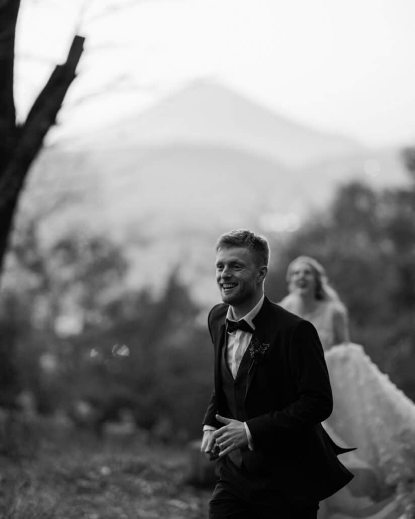 The groom walks ahead smiling, the bride laughing behind him, with a misty mountain peak beyond the trees on their Japan elopement.