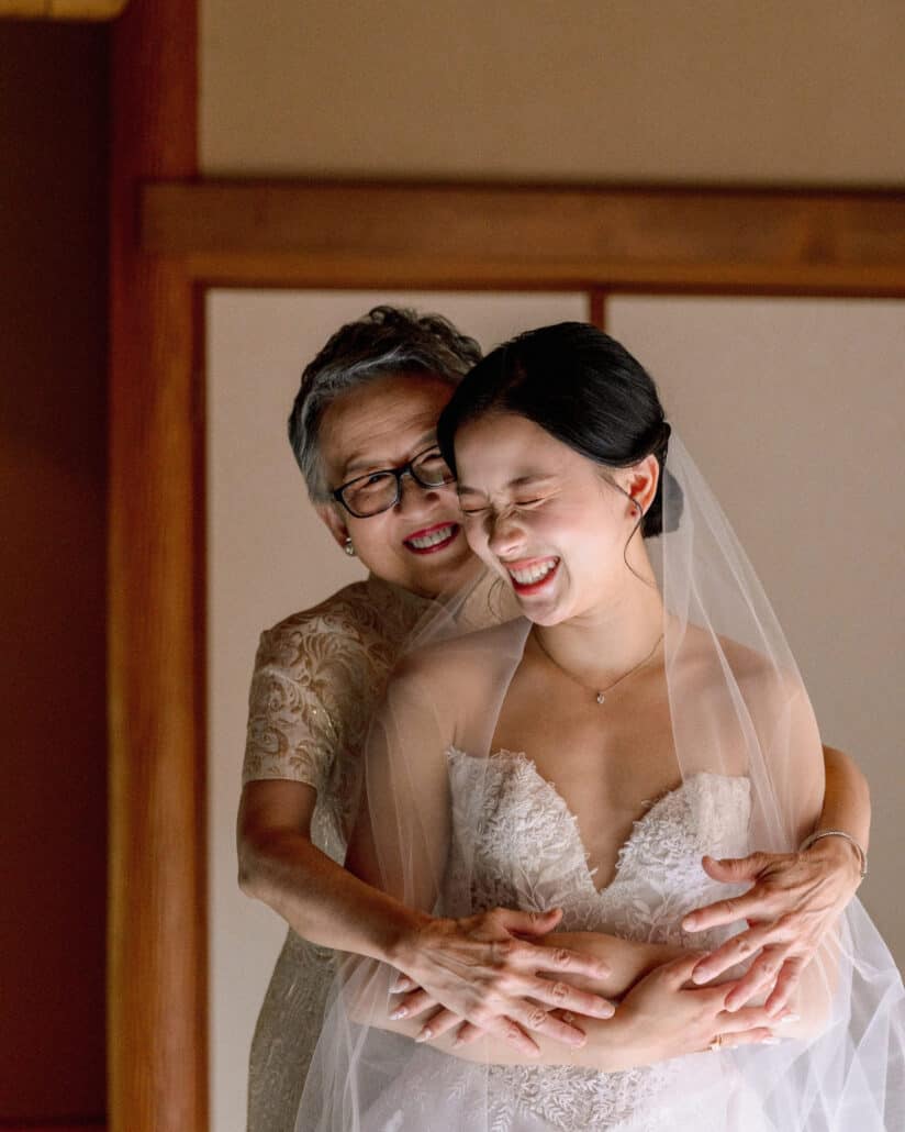 The bride's mother hugs her from behind as they both laugh, veil on, while the bride gets ready in a wood-framed ryokan room at Jinya.