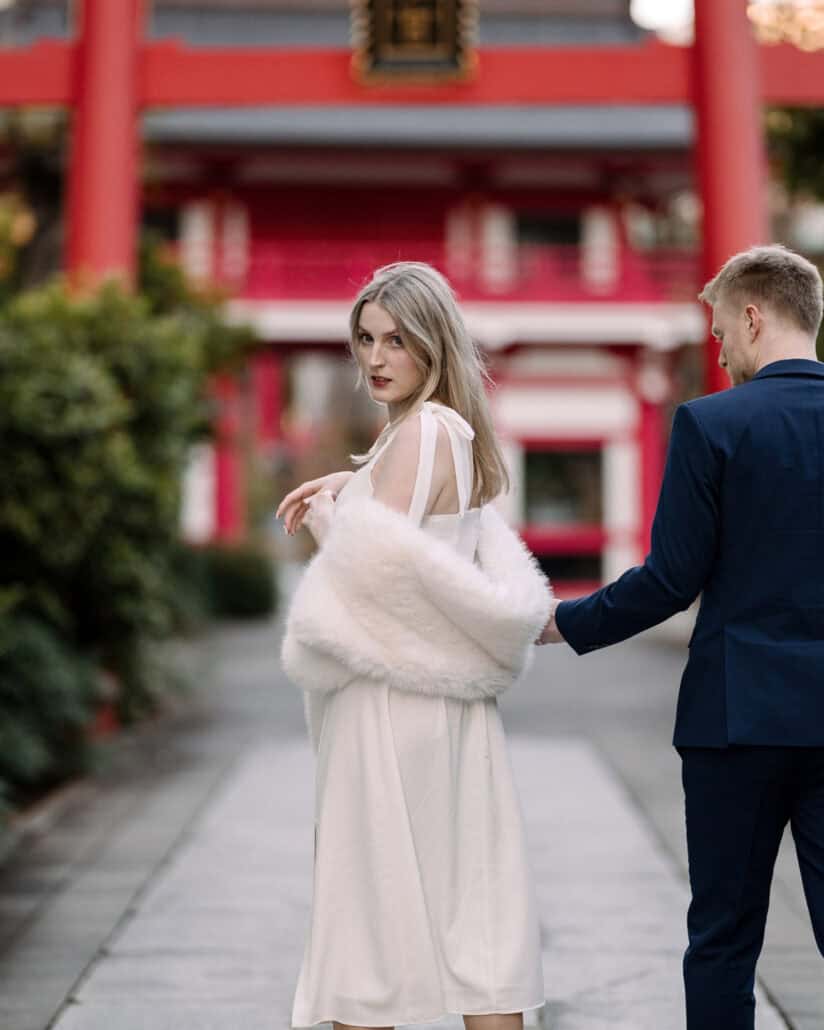 The bride looks back over her shoulder as the groom leads her along the stone path of a vermilion shrine, a Tokyo couple photoshoot.