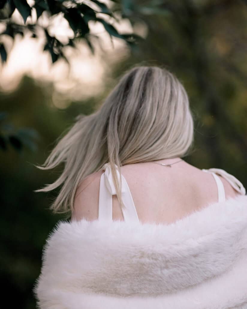 The bride turns away beneath dark leaves, her blonde hair swinging over a white fur stole, during a Tokyo pre wedding photoshoot.