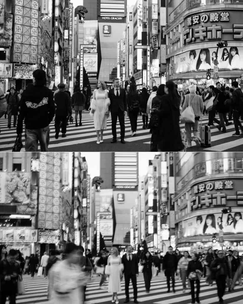 In black and white Shinjuku street photography, the couple pause mid crossing in Kabukicho as the crowd passes, the Godzilla head behind them.