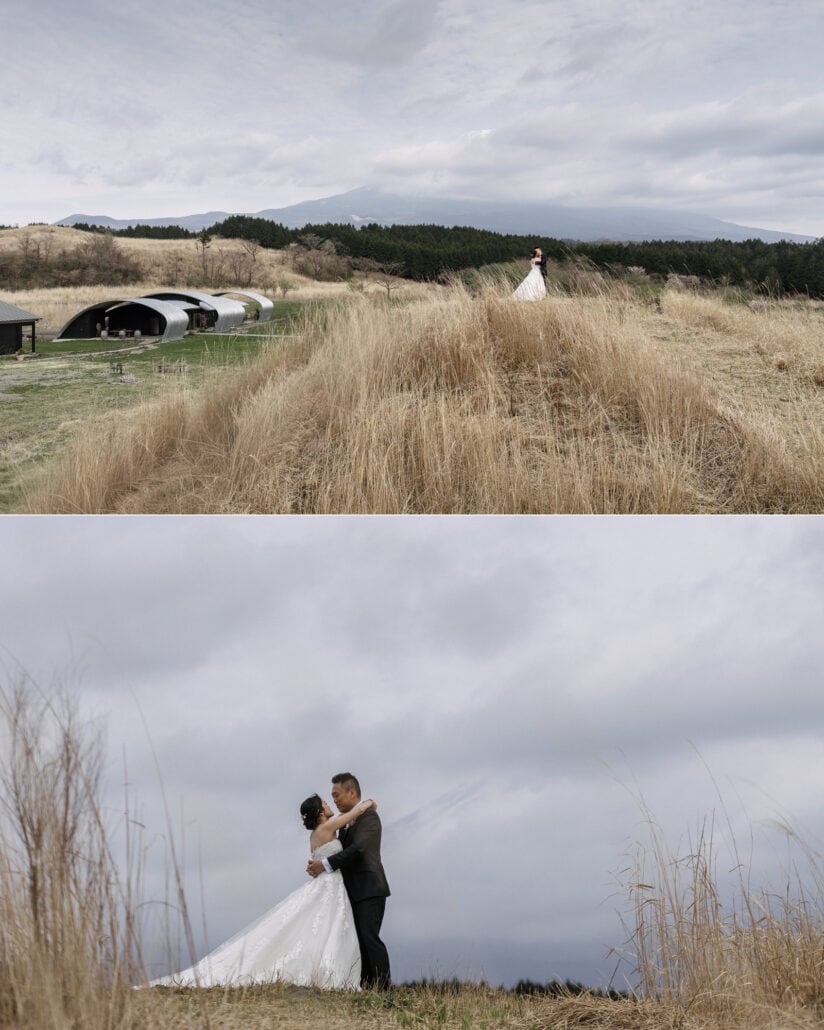 The couple stand small on a ridge of golden grass above the vaulted buildings of Fujisan Winery, then embrace in tall dry grass under a grey sky.