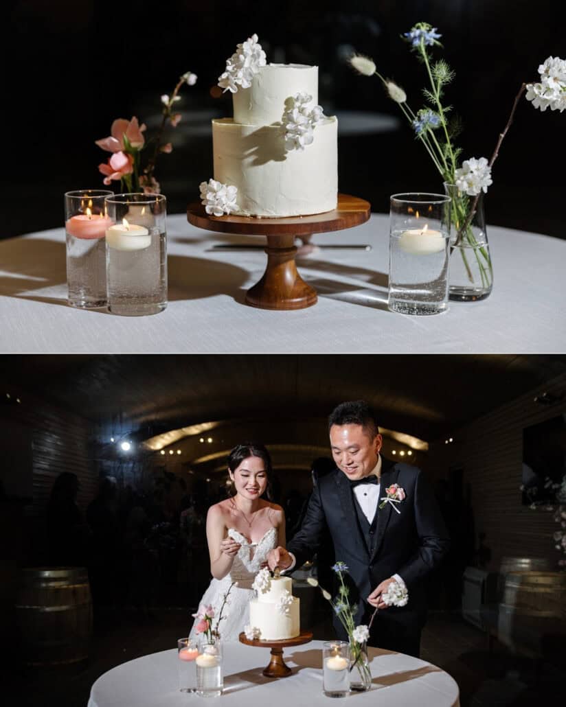 A two tier white cake with white flowers sits among floating candles and bud vases, and the couple cut it in the barrel room at Fujisan Winery.