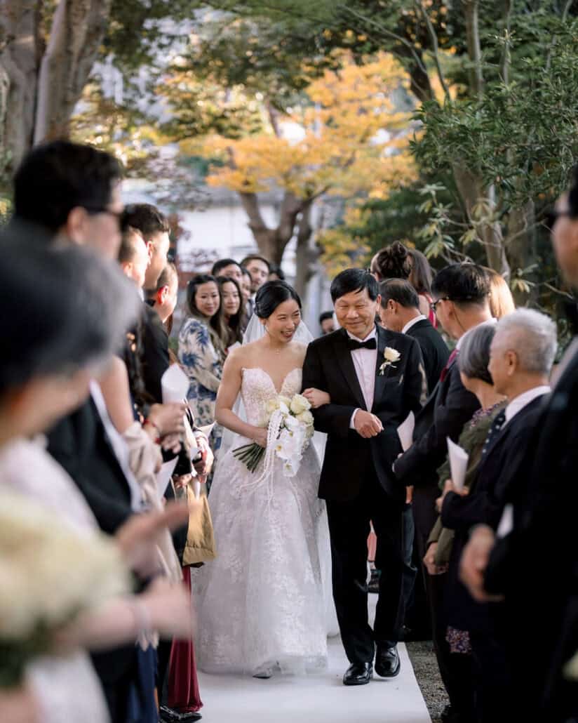 The bride walks down the white aisle at Jinya on her father's arm as guests stand on both sides, the processional of their Japanese garden wedding.