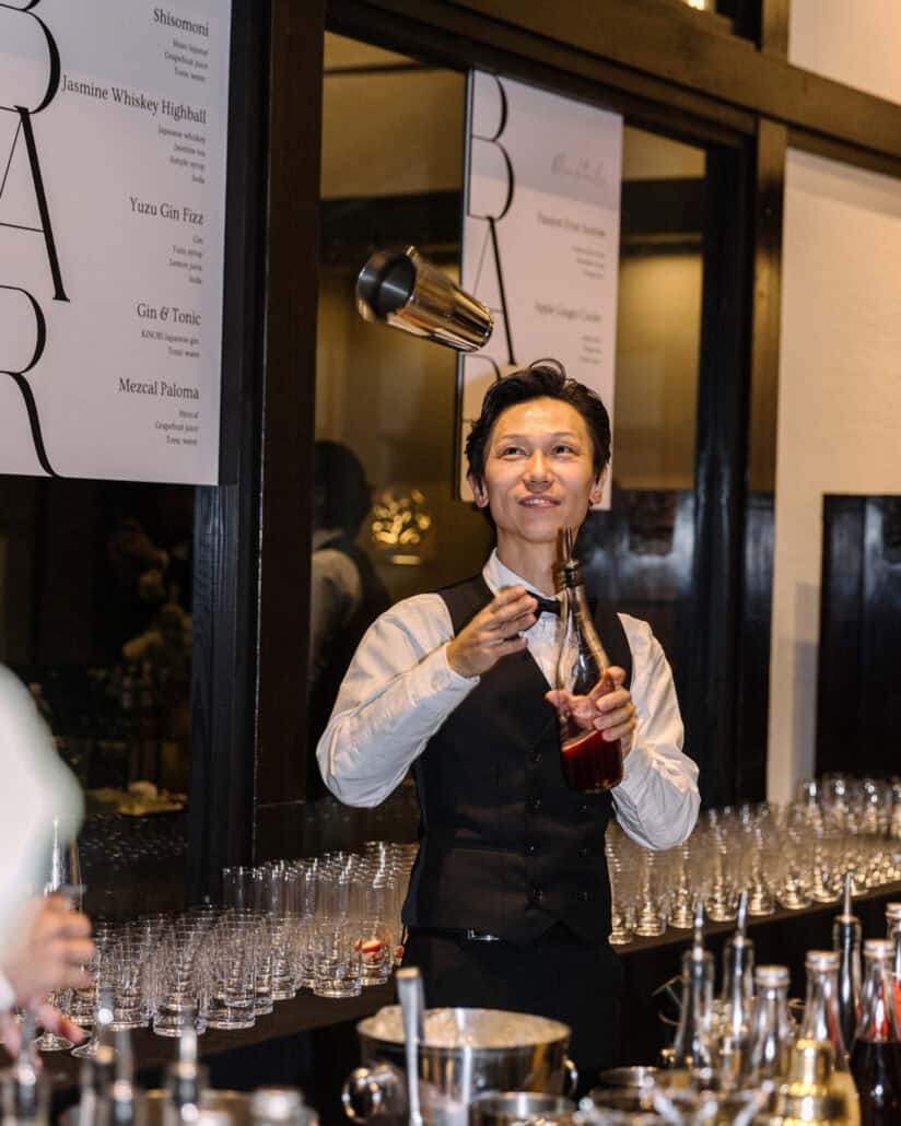 A flair bartender in a waistcoat and bow tie sends a shaker flying overhead at the wedding cocktail bar at Jinya, rows of glasses lined up below.
