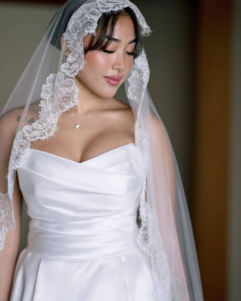The bride in a lace-edged mantilla veil and strapless gown, eyes lowered, getting ready for her cherry blossom wedding in Japan.