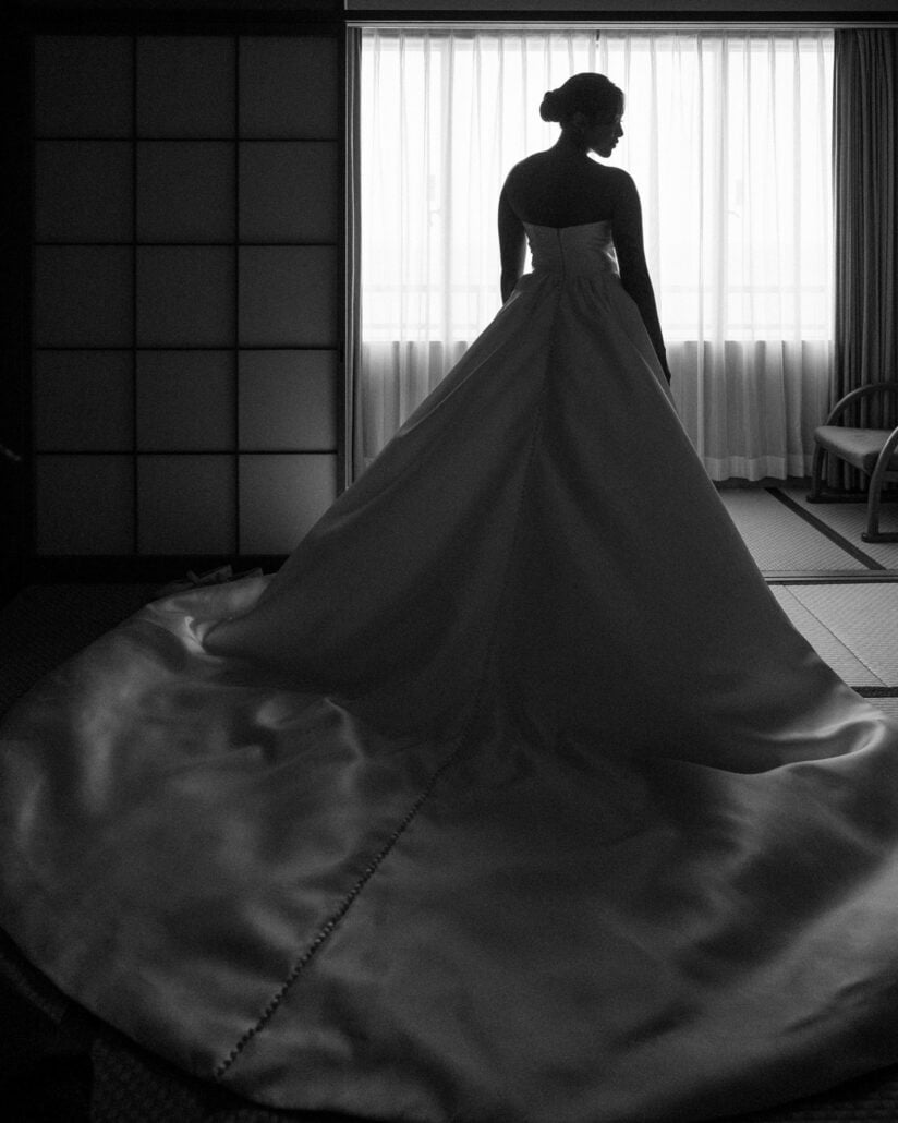 Black and white silhouette of the bride in her full-skirted gown at a curtained window in a tatami room with shoji screens.