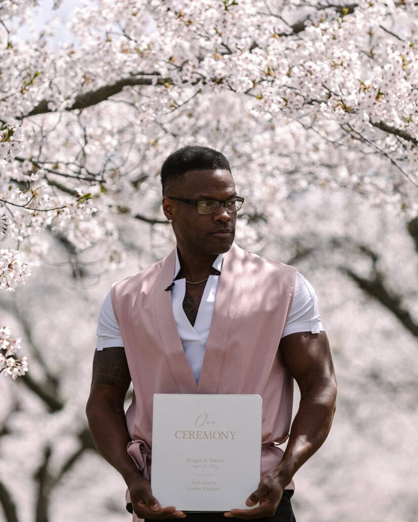 Their friend in a pink waistcoat holds the ceremony booklet under the cherry trees, ready to lead the cherry blossom wedding.