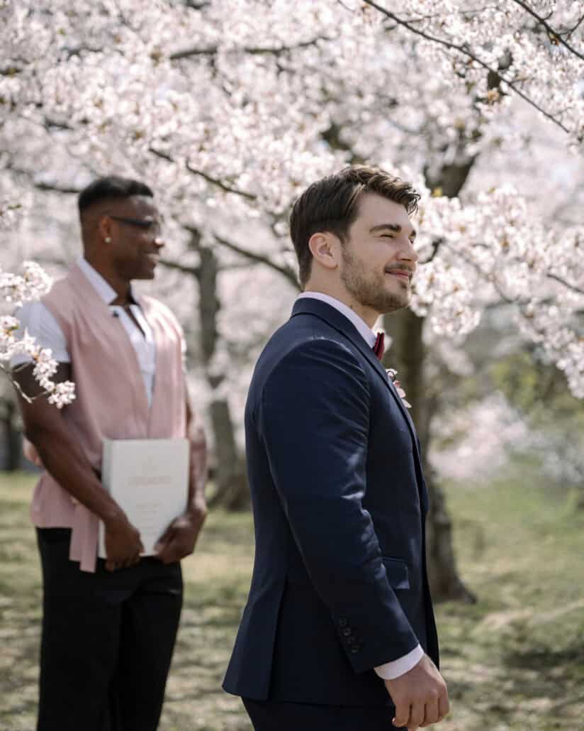 The groom in a navy suit and burgundy bow tie waits beside their friend and the ceremony booklet under the cherry trees.