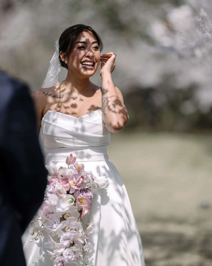 The bride wipes her eye and laughs at the same time during her cherry blossom wedding ceremony, dappled shade on her gown.