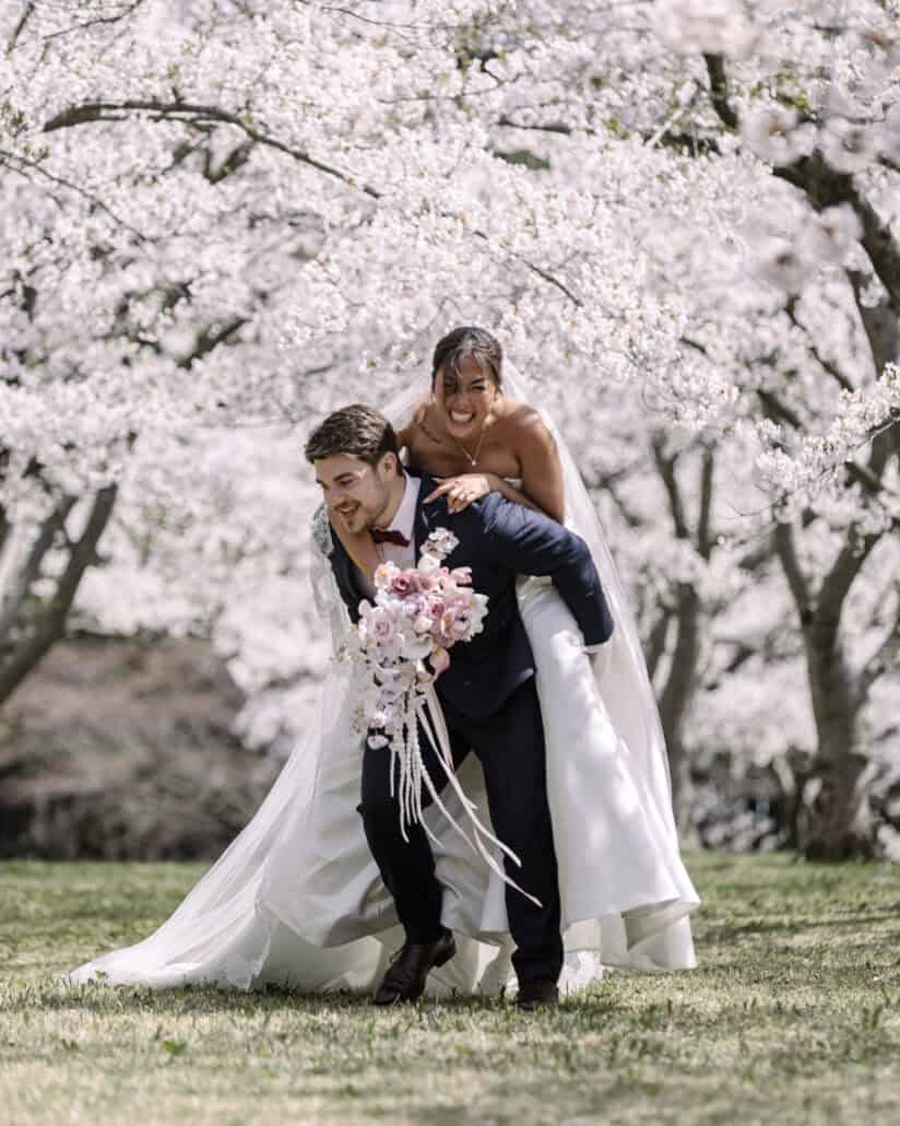 The groom gives the bride a piggyback under the cherry blossoms, her bouquet in one hand and a wide smile on her face.