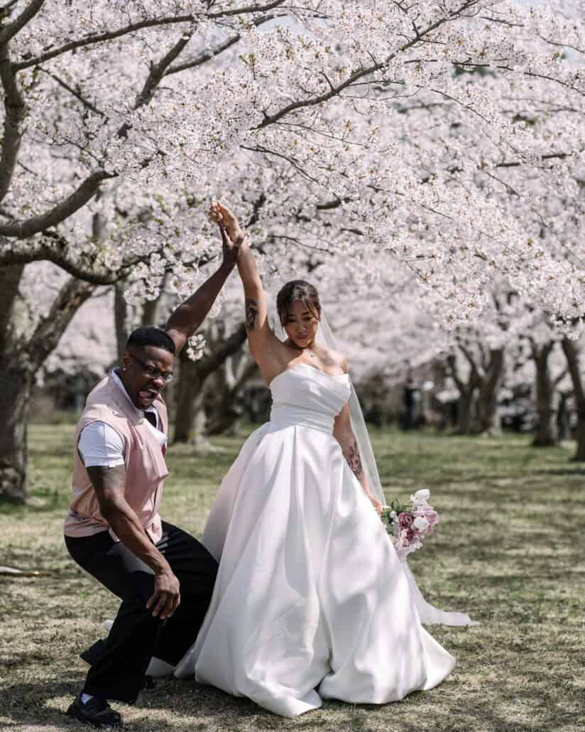 Their friend in a pink waistcoat and the bride throw their arms up together under the cherry blossoms after the ceremony.