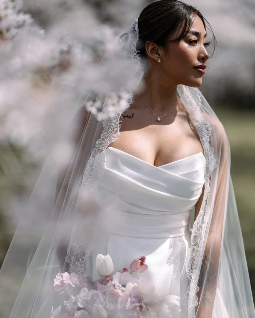 A profile portrait of the bride in her lace mantilla veil, blurred cherry blossoms in the foreground at her wedding in Aomori.