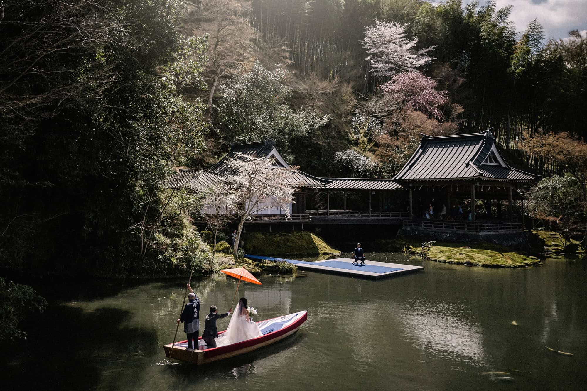 The bride crosses the pond by boat under a red paper umbrella towards the Noh stage at Asaba Ryokan, with cherry blossom and bamboo on the hill behind.