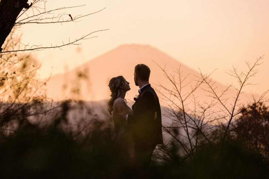 The bride looks up at the groom as he holds her, with Mt Fuji behind them in a pink sunset haze, at the end of an autumn elopement in Japan.