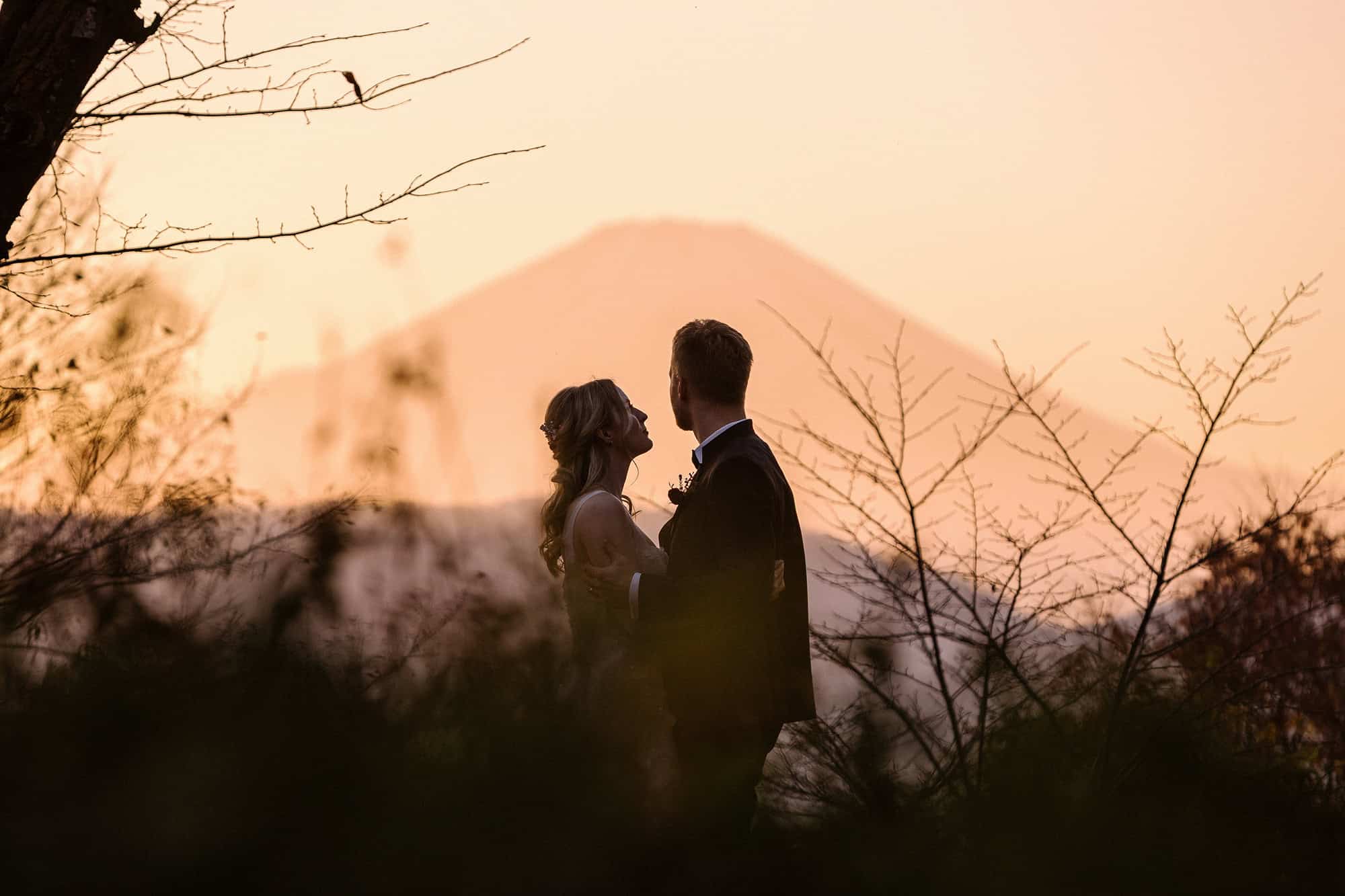 The bride looks up at the groom as he holds her, with Mt Fuji behind them in a pink sunset haze, at the end of an autumn elopement in Japan.