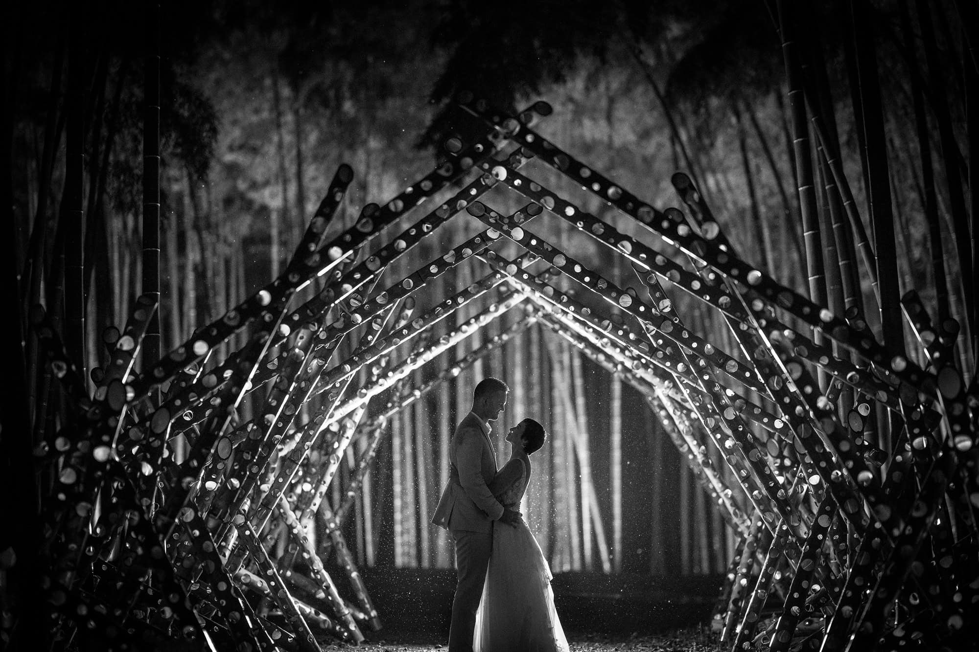 Silhouetted in the rain under a tunnel of drilled bamboo lit from inside, the couple face each other in the Wakayama Farm bamboo forest at night.