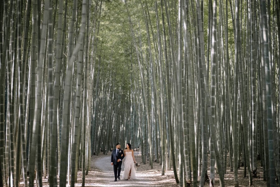 The bride and groom walk arm in arm up a pale path between walls of tall bamboo at Wakayama Farm, a bamboo forest near Tokyo.