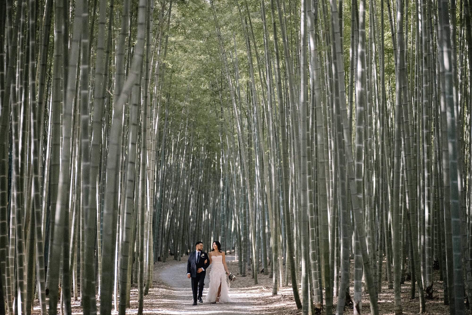 The bride and groom walk arm in arm up a pale path between walls of tall bamboo at Wakayama Farm, a bamboo forest near Tokyo.