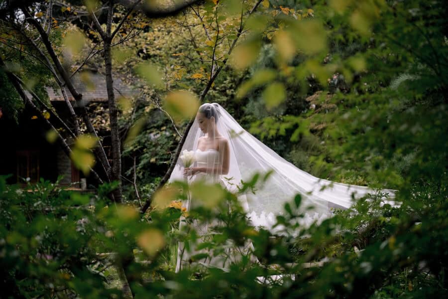 The bride, in a long veil edged with lace, holds white calla lilies in a ryokan garden framed by leaves, at a Greek and Japanese wedding in Japan.