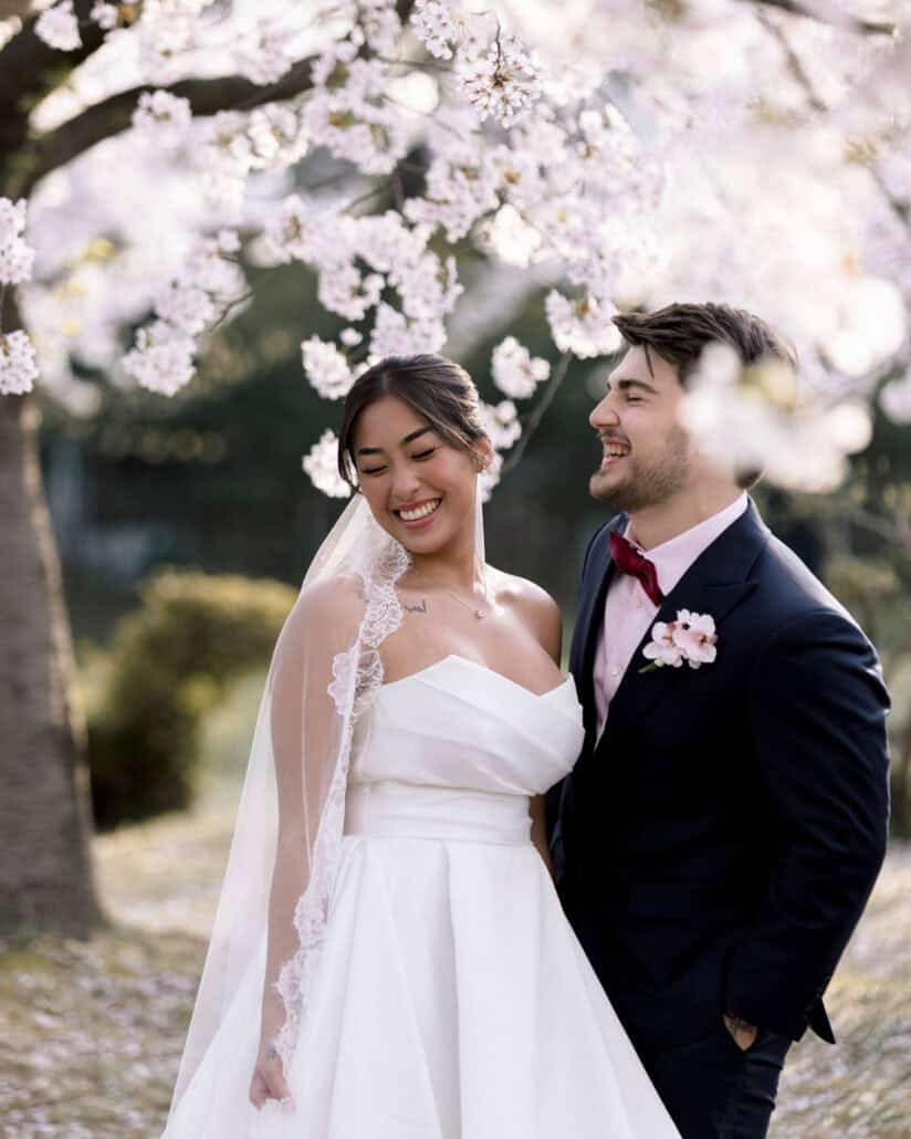 The couple laugh together under a branch of white cherry blossoms in Hirosaki, her lace veil catching the light.