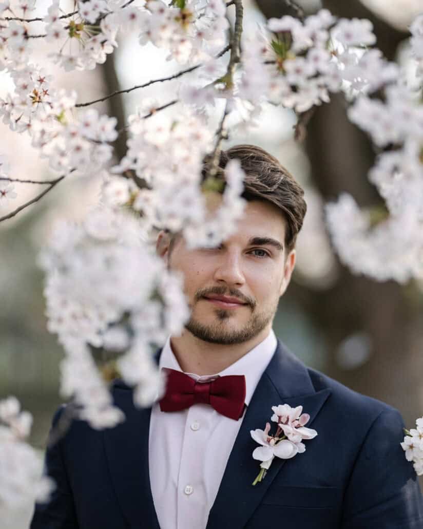 A portrait of the groom through the cherry blossoms of Hirosaki, his burgundy bow tie and pink orchid boutonniere in view.