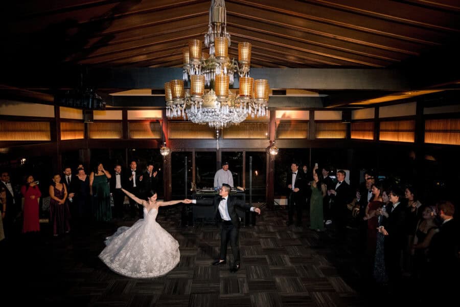 The bride twirls in her first dance under a chandelier as guests circle the floor of a wood-beamed hall at Jinya, a moment their wedding film keeps.