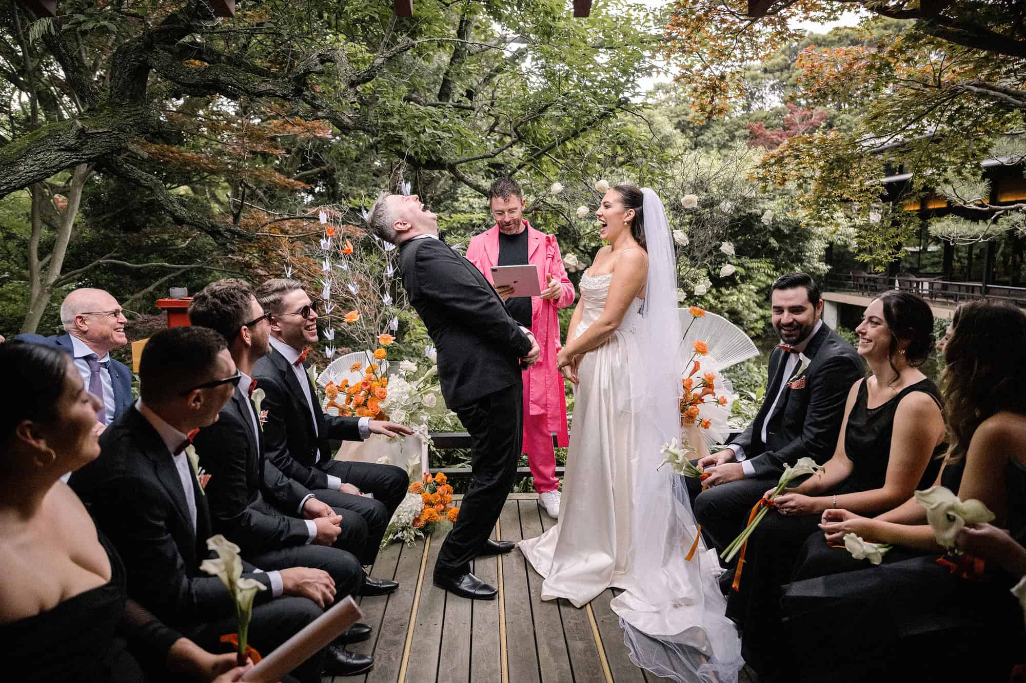 The groom laughs with his head thrown back beside the bride and the officiant in pink at a June wedding in Japan, on the garden deck at Jinya, Hadano.