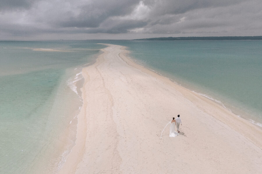 The couple walk a long sandbar through turquoise sea under storm cloud, her veil trailing, the day before their Rosewood Miyakojima wedding.