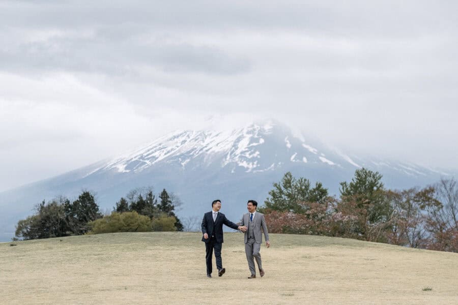 Mt Fuji engagement photos near Kawaguchiko, the couple laughing hand in hand across a grassy rise as the snowy summit hides in cloud behind.